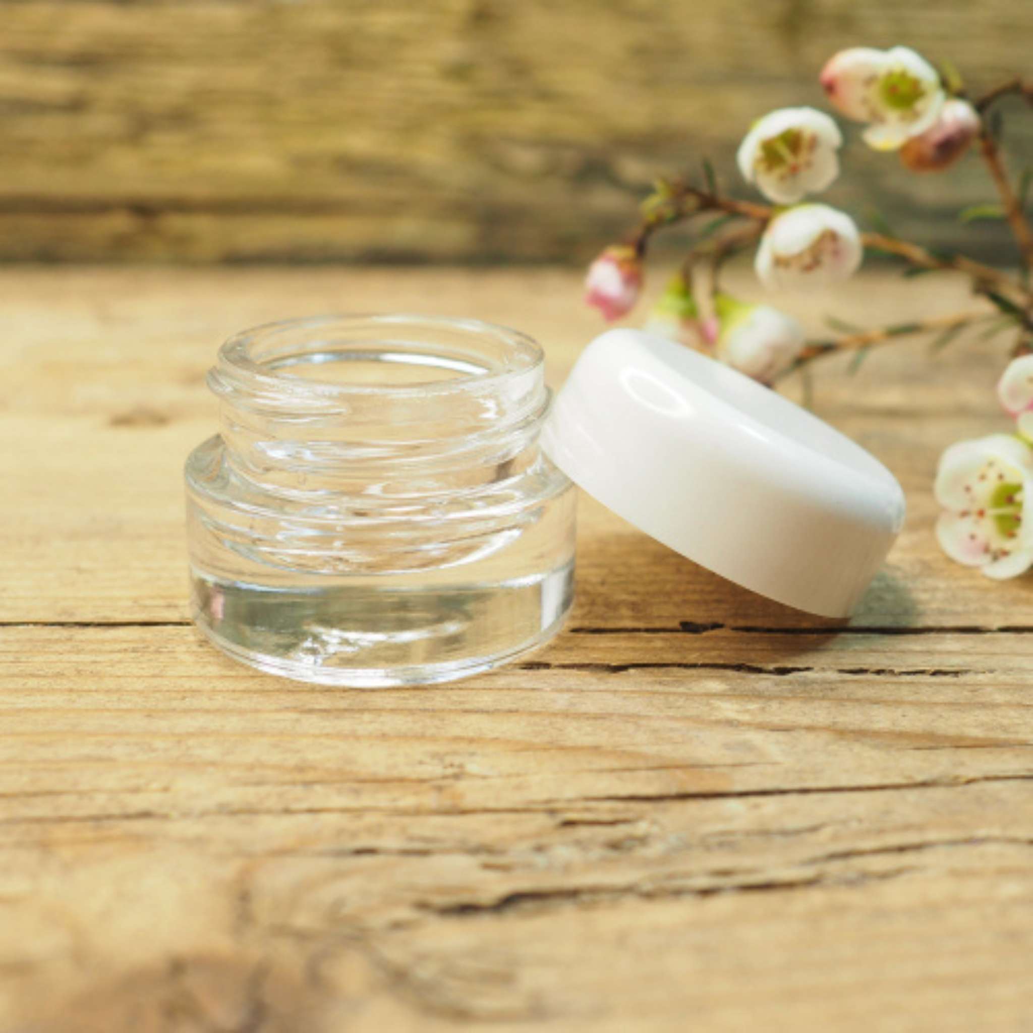 transparenter Glastiegel mit weißem Deckel, geöffnet, auf rustikalem Holzuntergrund, florale Dekoration im Hintergrund