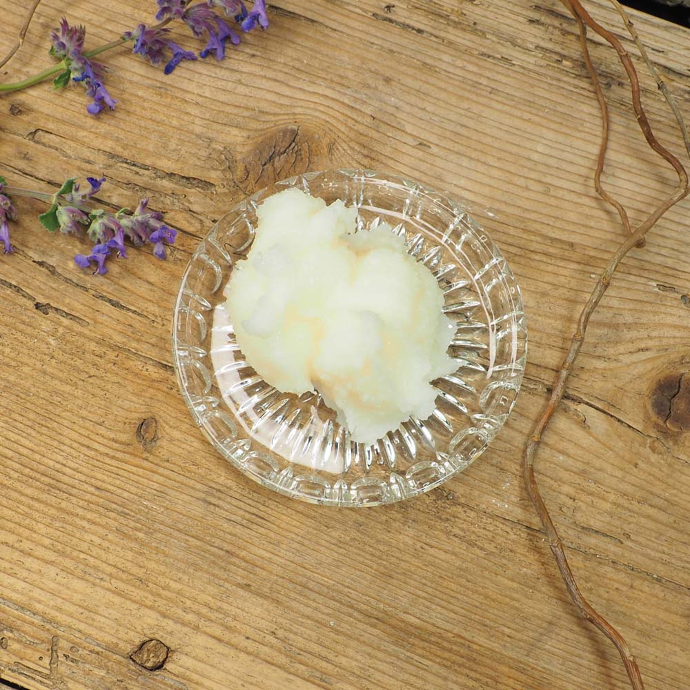 Portion Olivenbutter auf einem Glas-Teller, umgeben von lila Blüten und einem Zweig auf einem rustikalen Holztisch