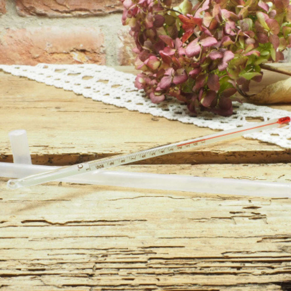 Klassisches Glasthermometer mit roter Spitze auf rustikalem Holzuntergrund mit Blüten im Hintergrund.